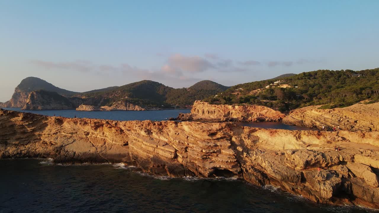 Ripresa aerea con zoom in durante il tramonto del paesaggio roccioso e montuoso su una spiaggia a Ibiza, Spagna con una barca e turisti