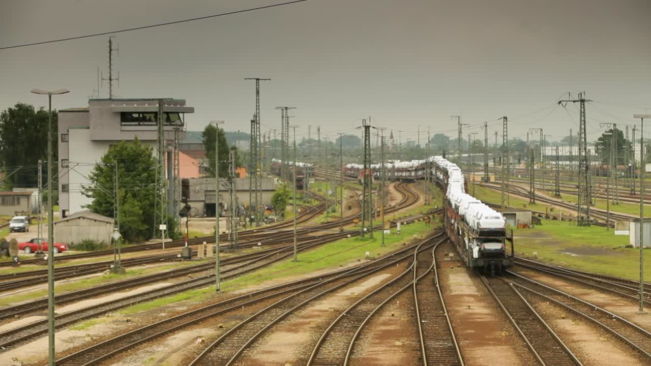 높은 시야에서  ⁇ 드헬드 카메라가 하 ⁇  화물차로 가득 찬 열차로  ⁇ 비는 기차 야드를 포착합니다. 왼쪽에 있는 컨트롤 타워는 구름이 가득한 하늘 아래 푸른 나무로 둘러싸여 있습니다.