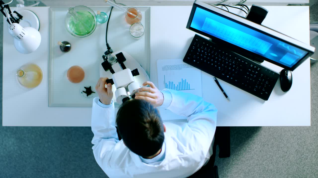 초록색 화면이 켜진 개인용 컴퓨터에서 일하는 과학자가 현미경을 들여다보고 차트를 가진 문서에 결과를 적어 놓는 위쪽.