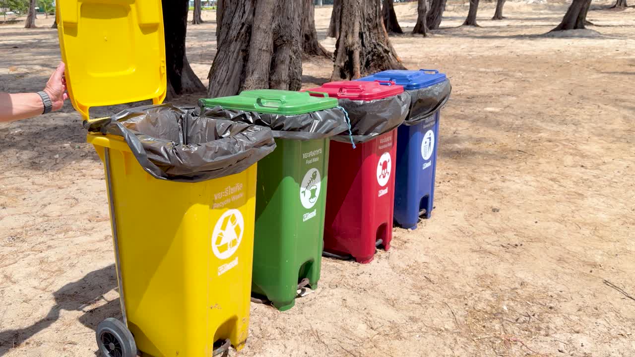 A hand closes the lid of a yellow recycling bin outdoors on sandy ground