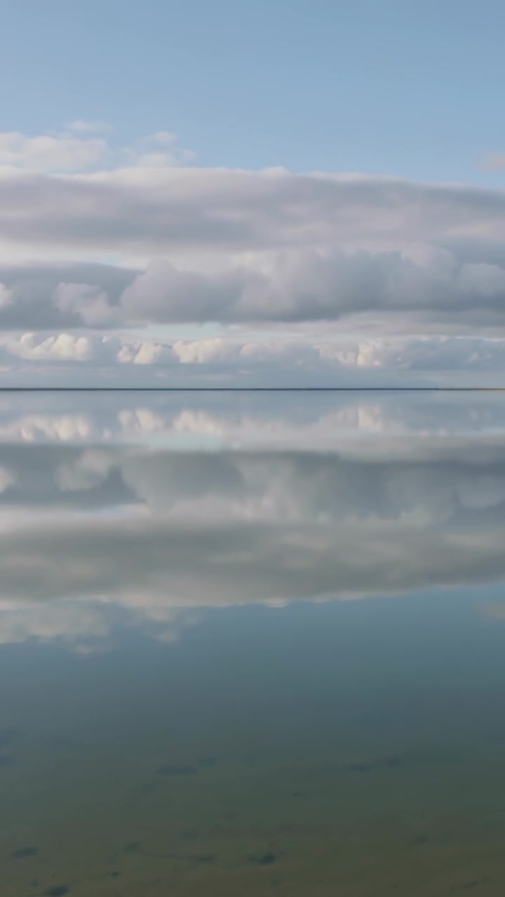 Video verticale: Telecamera che si inclina rivelando cielo e acqua che riflettono le nuvole sul fondo sabbioso di un lago poco profondo