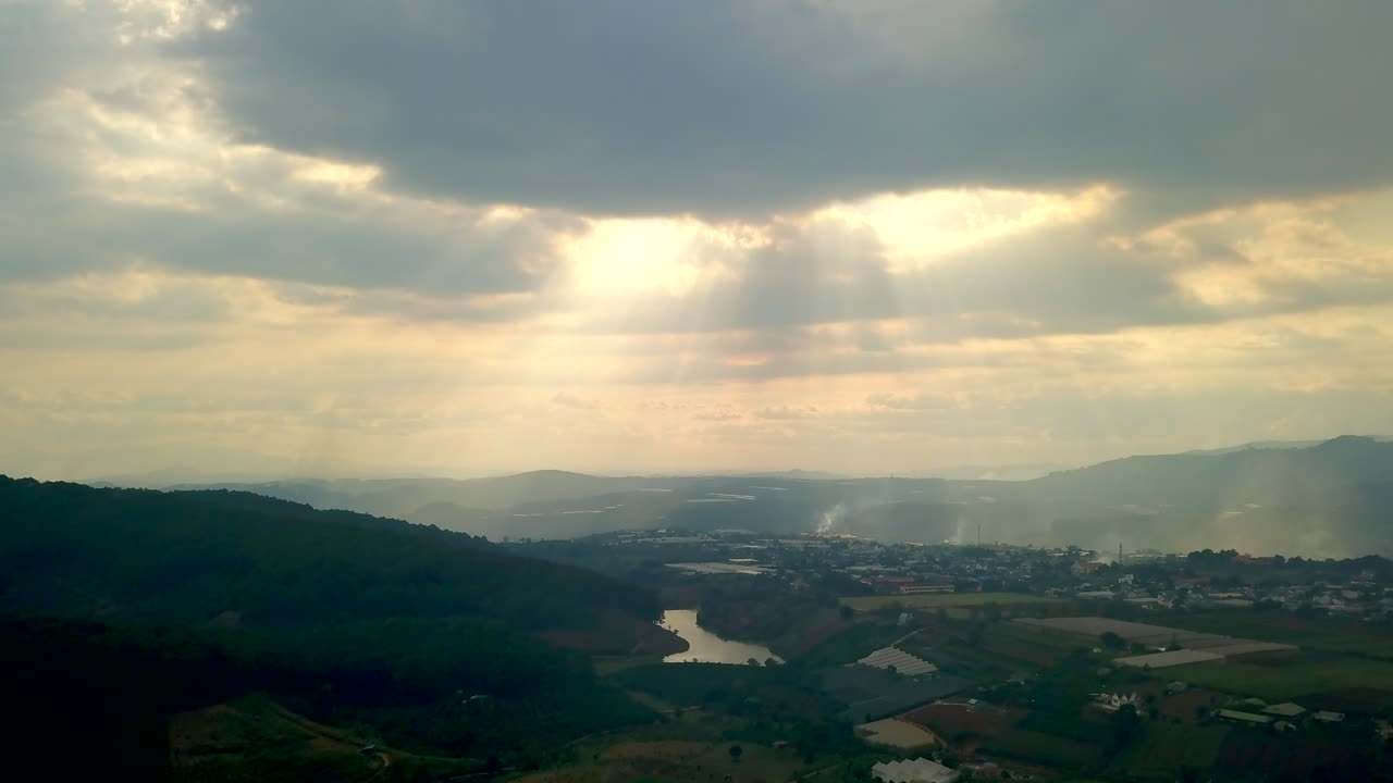 Drone shot of the sun shining through clouds with beautiful sun rays showing over hills of Dalat, Vietnam in Asia. Green forest and mountains and village.