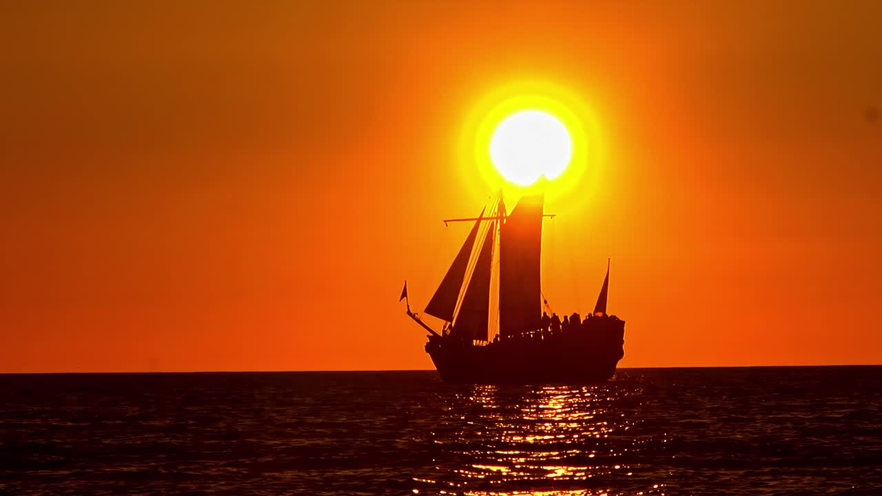 A beautiful slow-motion shot shows the silhouette of a vintage tall ship with tourists sailing peacefully across the Baltic Sea as the sun sets on the horizon, creating a golden glow