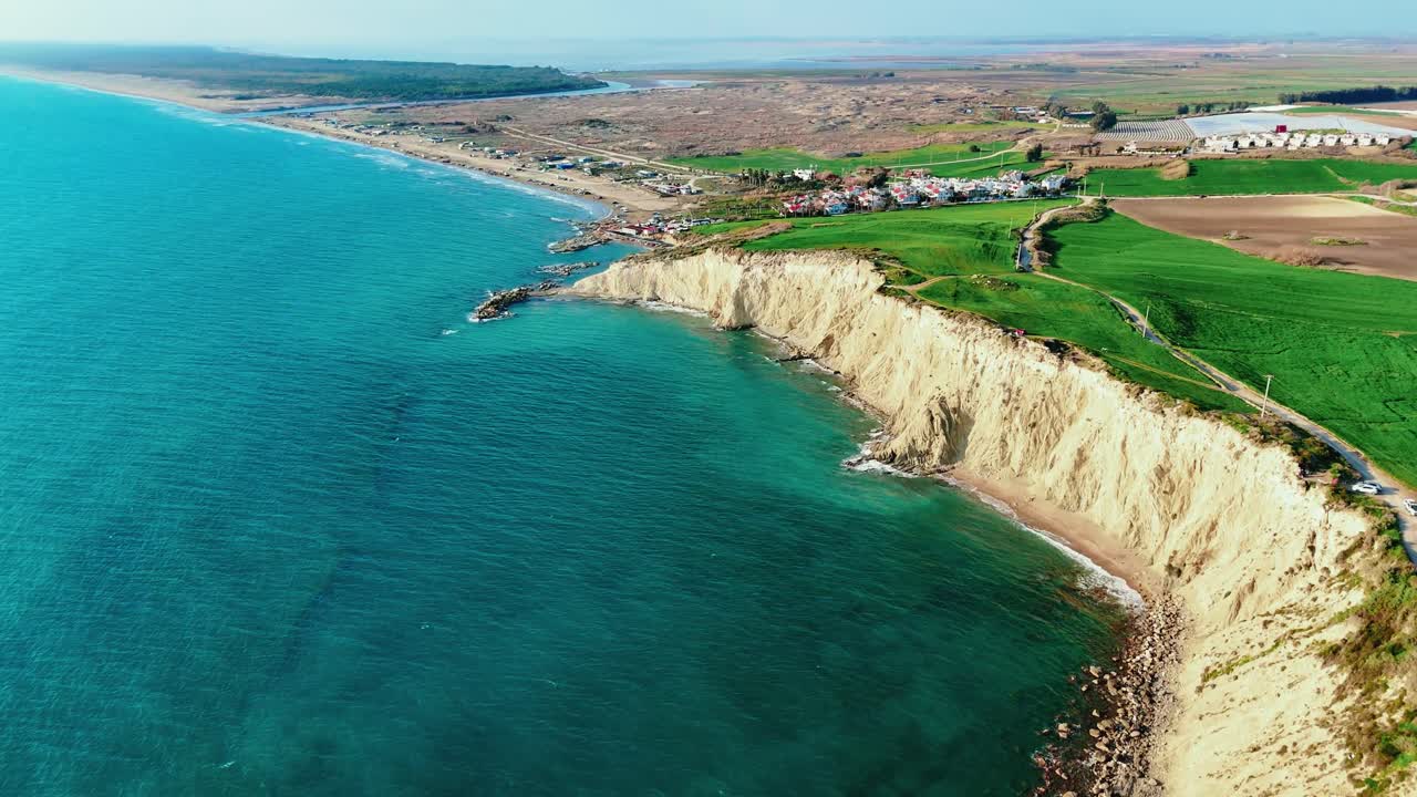 Aerial view of rugged coastal cliffs dropping into a clear turquoise sea, surrounded by lush green farmland and distant villages, showcasing the diverse natural landscape in Vietnam