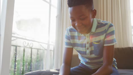 Feliz-Niño-Afroamericano-Sentado-En-El-Sofá-Usando-Un-Teléfono-Inteligente-Y-Sonriendo,-Iluminado-Por-El-Sol