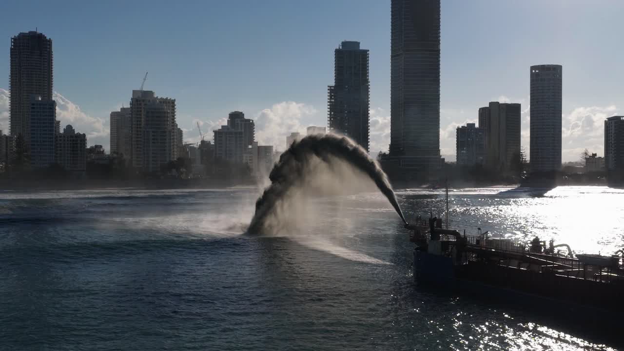 A dredging ship pumps sand onto a beach, silhouetted against a city skyline, under bright daylight