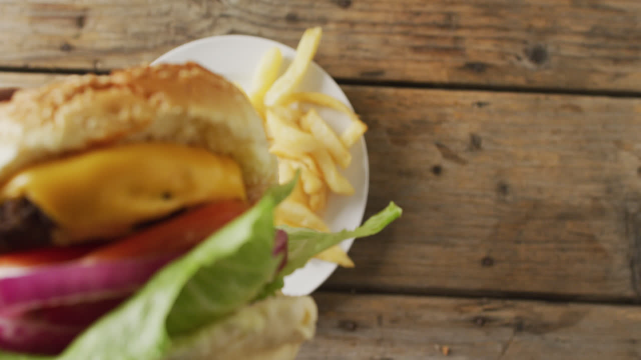 video de pov de la mano afroamericana sosteniendo una hamburguesa con queso, sobre patatas fritas en un plato en una mesa de madera