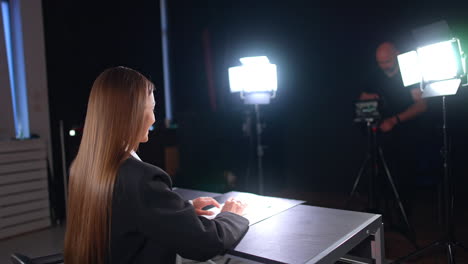 positive brunette woman with long straight hair sits at the desk half-back to camera. lights of soffits illuminate the blogger recording a video. male photographer at backdrop.