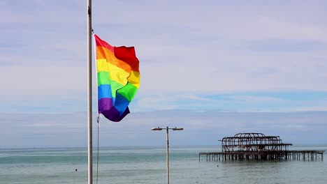 the rainbow flag, with its six colours fluttering in the breeze on a sunny day in brighton with the burnt out west pier in the background