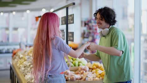 Un-Chico-Moreno-Feliz-Con-Auriculares-Blancos-Baila-Con-Su-Novia-De-Pelo-Rosa-Durante-Una-Compra-General-En-Un-Supermercado-Cerca-De-Los-Estantes