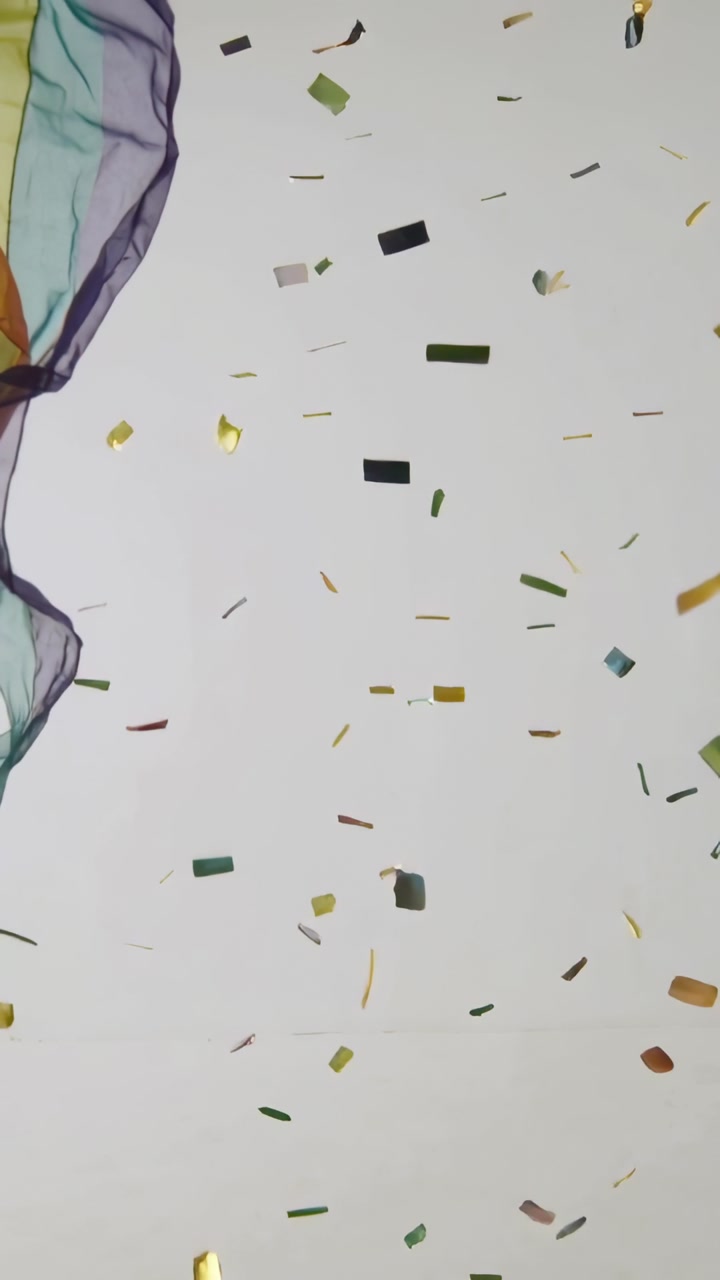 Vertical video: Falling foil bits shining at pale wall by gravity and air, sheer scarves left right