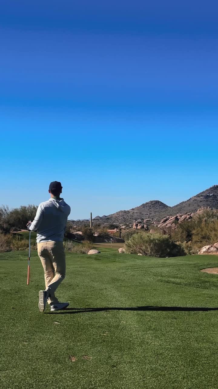 Golfer hits iron with a draw into protected green on pristine Arizona desert golf course, cactus and mountain backdrop, windy, vertical 4k sunny day slow motion