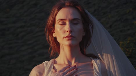 at sunset, a woman wearing a white dress and veil meditates with her eyes closed and her hand on her chest, embracing spirituality and mindfulness