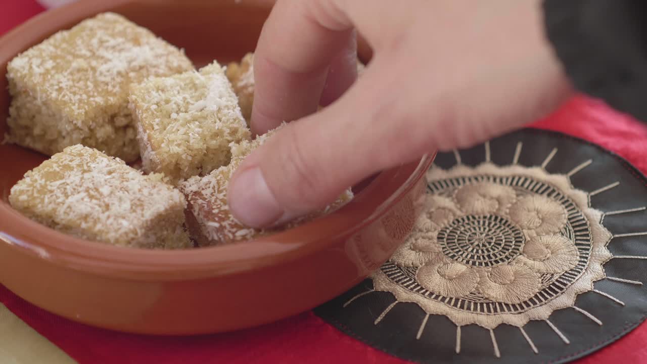 En hånd tager sunde hjemmelavede kokosbidder eller -kage fra en traditionel terrakotta skål på en blondebrik. Naturligt lys fremhæver præsentationen af den økologiske vegetariske snack.