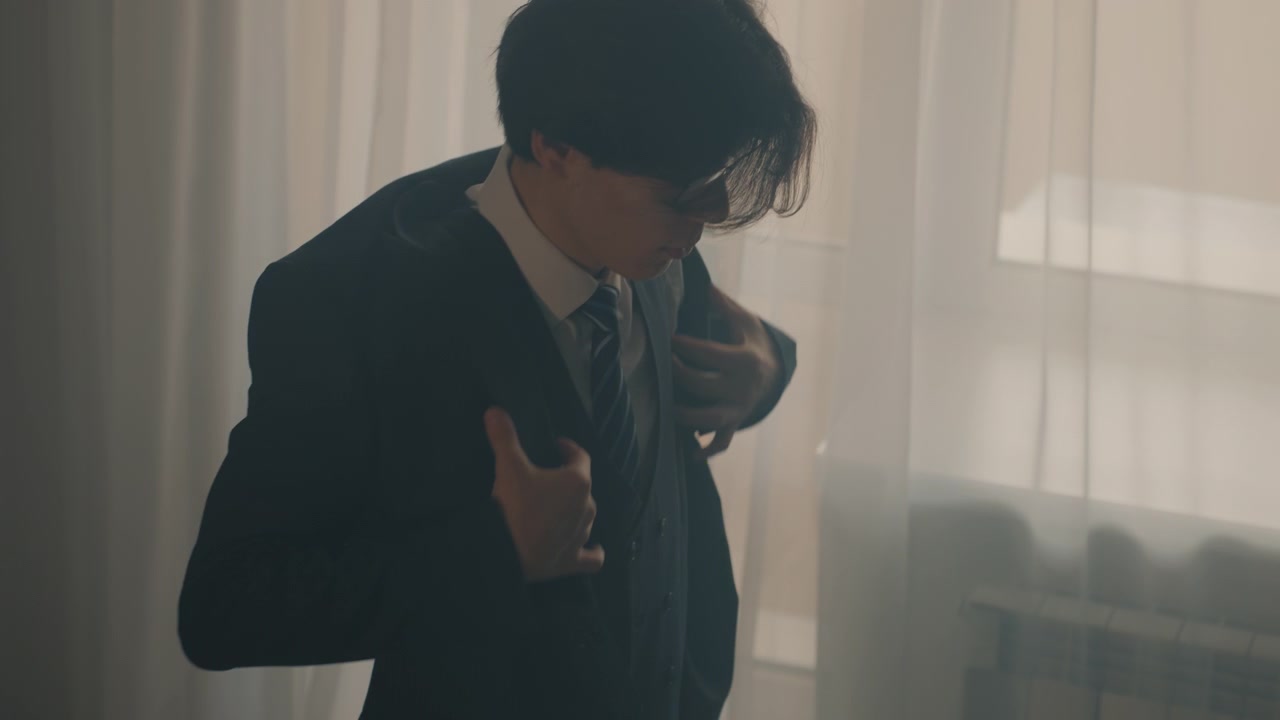窓際でジャケットを羽織り、集中した緊張した表情でネクタイを直すアジア人学生。薄手のカーテン越しに差し込む柔らかな逆光、椅子とベストが置かれたミニマリストな部屋、温かみのあるフィルム調のトーン、親密な雰囲気。