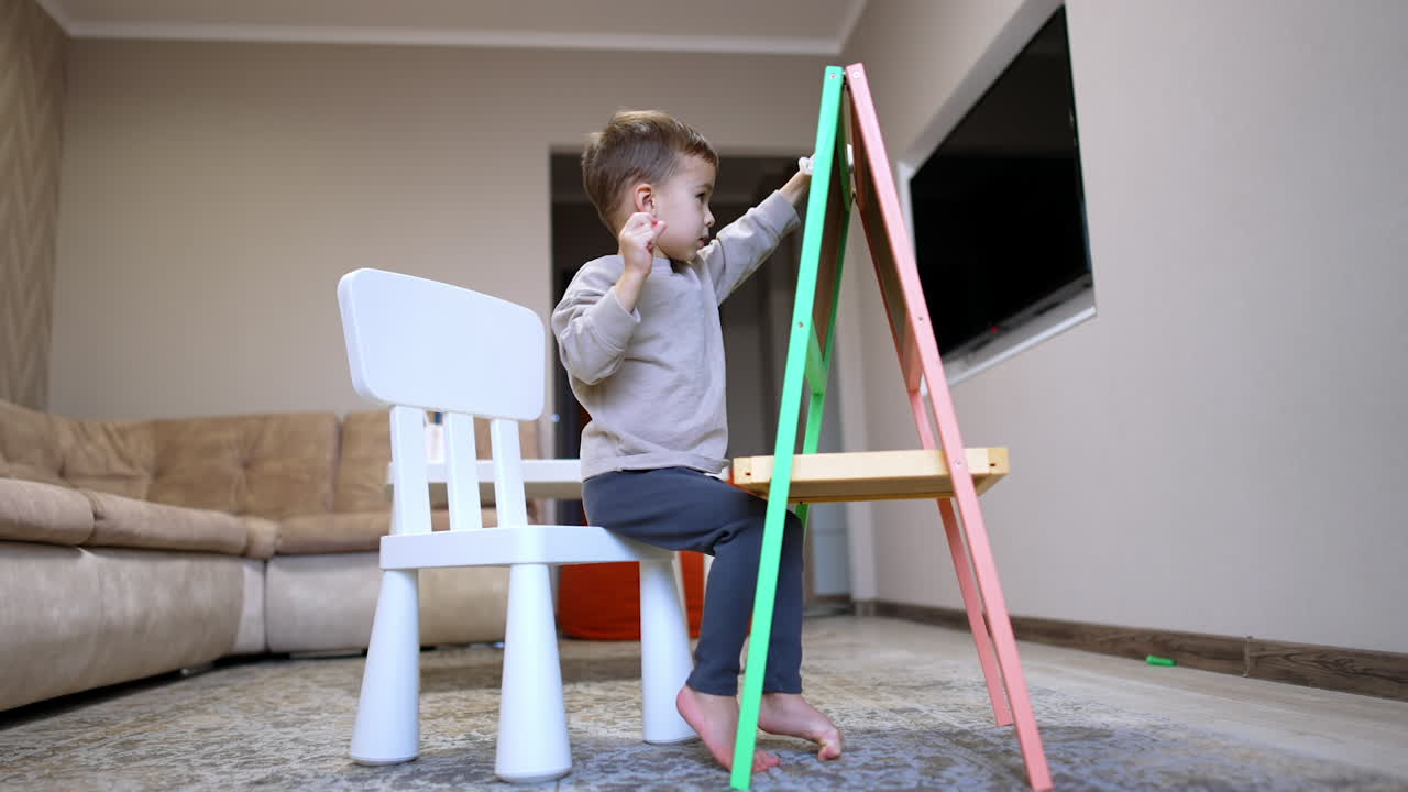 Young child drawing creatively at home. A small child sits at a chair, focused on drawing on an easel in a cozy living room during the day
