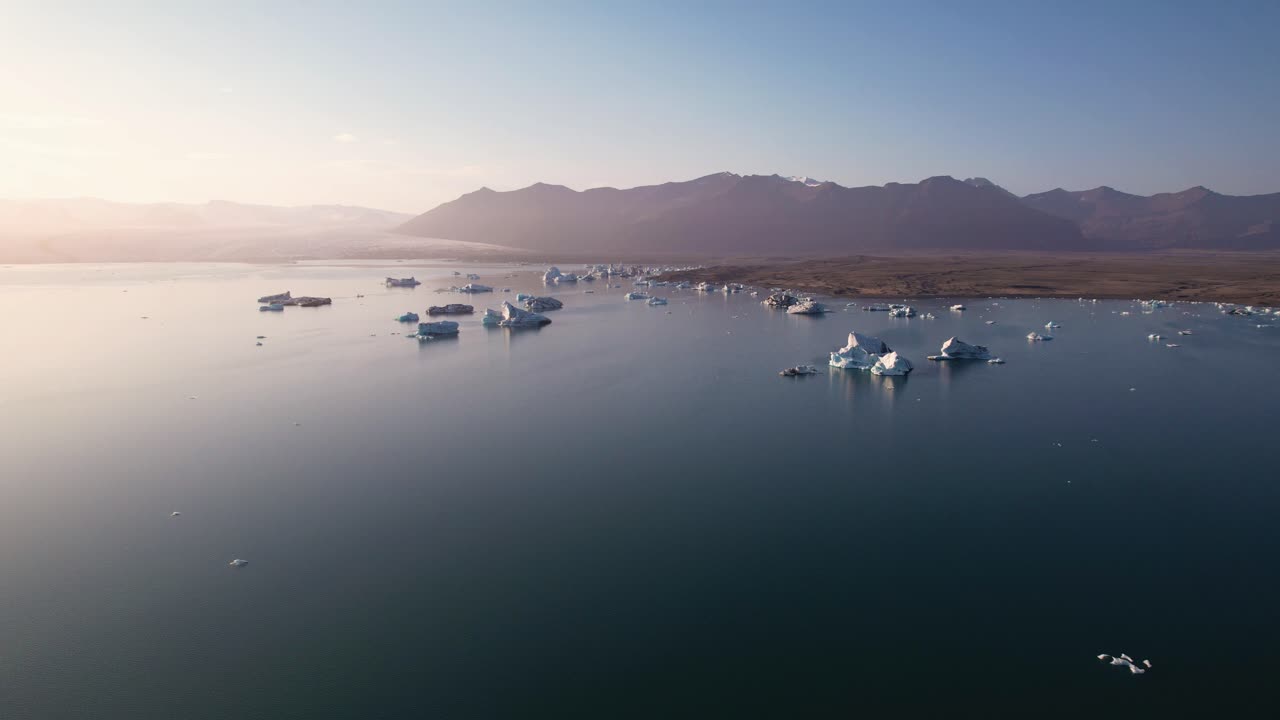 icebergs flotando en aguas tranquilas al amanecer con montañas en el fondo, sereno y majestuoso desde la vista aérea en islandia