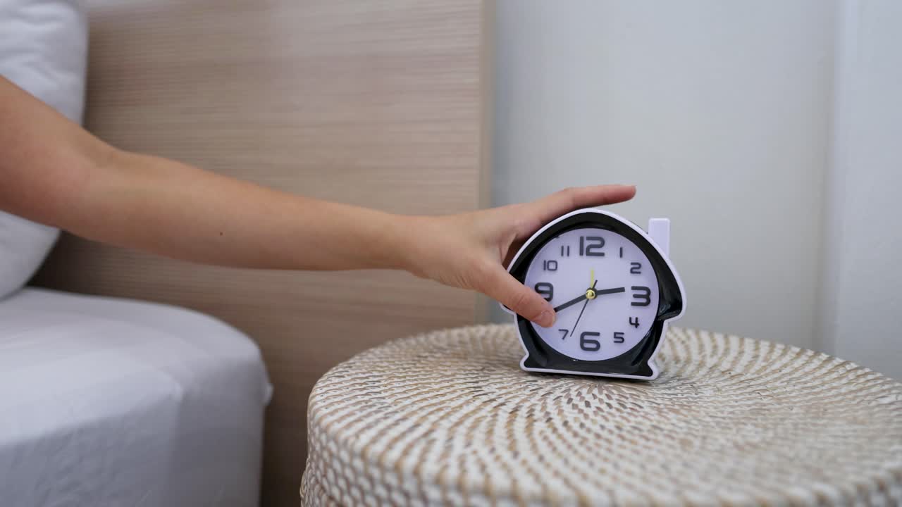 Una persona si allunga dal letto per spegnere una sveglia nera su un comodino