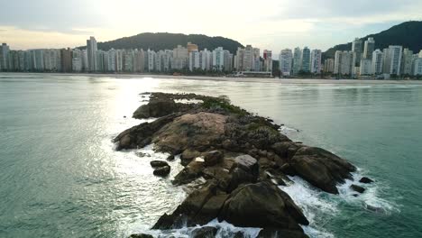 Drone-plane-approaching-a-small-rock-island-in-calm-and-peaceful-sea,-with-waves-crashing-on-the-rocks,-guarujá,-Brazil-beach