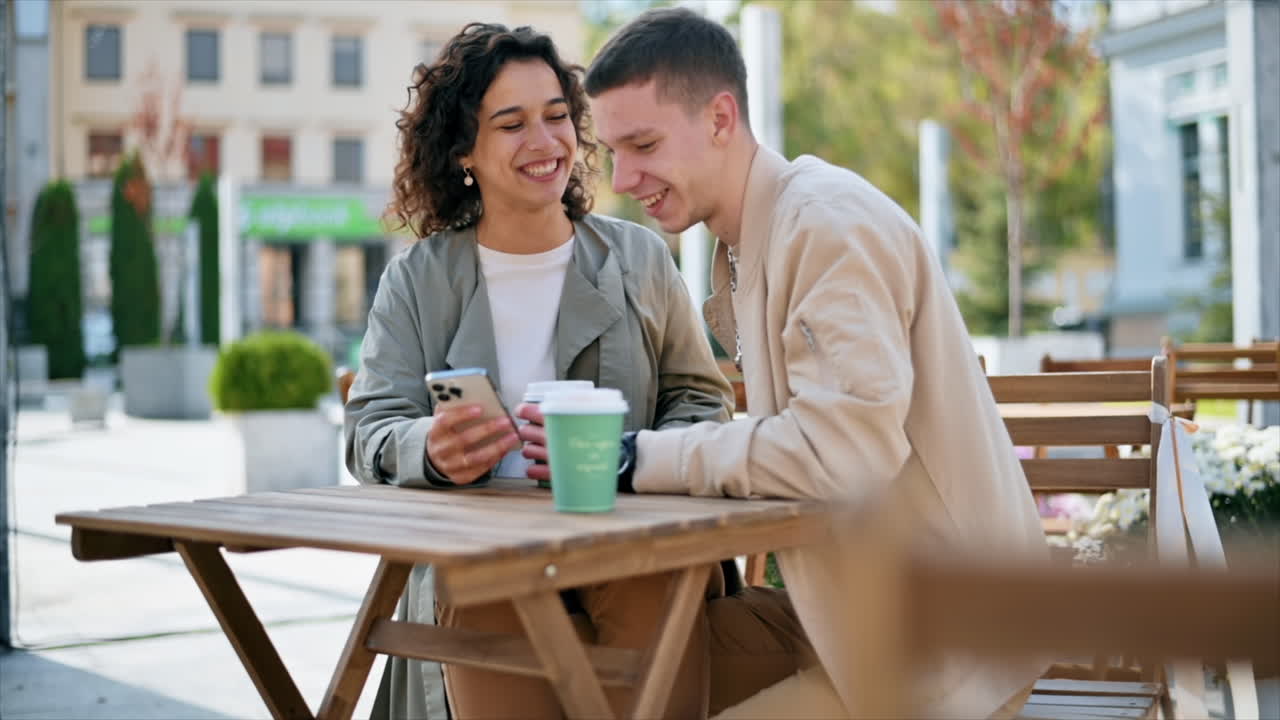 Una pareja feliz al aire libre cerca de una cafetería. Mirando el teléfono, riendo, hablando, café. Ambiente otoñal. Cámara lenta.