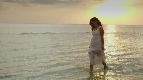 A-Young-Woman-In-A-Light-Summer-Dress-Is-Walking-Along-The-Edge-Of-The-Water-Against-The-Background-