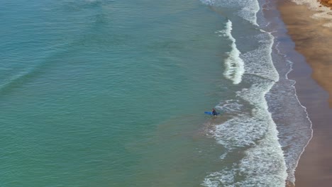 Drohnenaufnahme-Eines-Surfers,-Der-Aufs-Meer-Hinausgeht-Und-Auf-Ein-Surfbrett-Springt