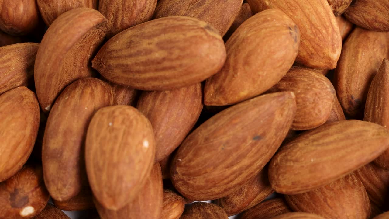Macro video of almonds falling onto a surface, creating a textured pile. Bright lighting enhances the natural brown tones