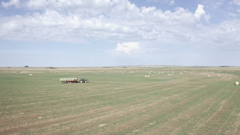 scenic flight above green farmland by farmer driving industrial farming tractor machine hauling bales of hay in flat expansive rural countryside and big sky, saskatchewan, canada, circle aerial