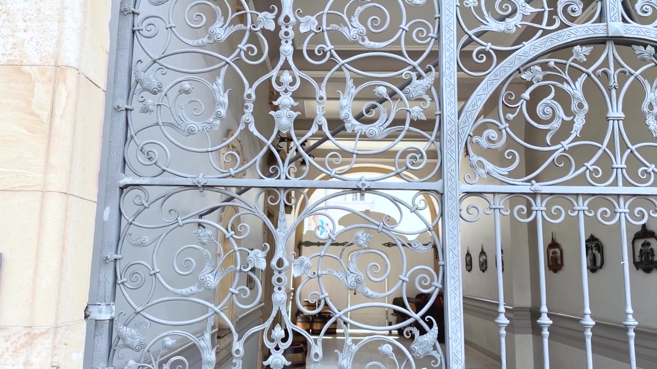 Un lent mouvement de caméra ascendant suit un portail d'entrée orné en fer forgé, révélant des volutes complexes et des détails métalliques artisanaux. Prise de vue en panoramique vertical ascendant filmée de près.
