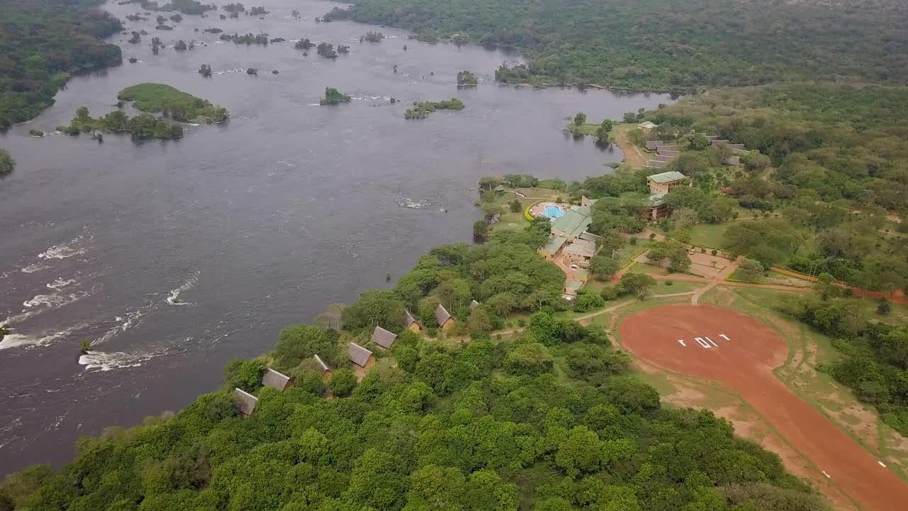 Complesso di lodge safari di lusso con una pista di atterraggio lungo il fiume Nilo, circondato da fitta boscaglia e isole fluviali nel Parco Nazionale delle Cascate Murchison, Uganda. Ripresa con drone che si allontana e rivela il paesaggio.