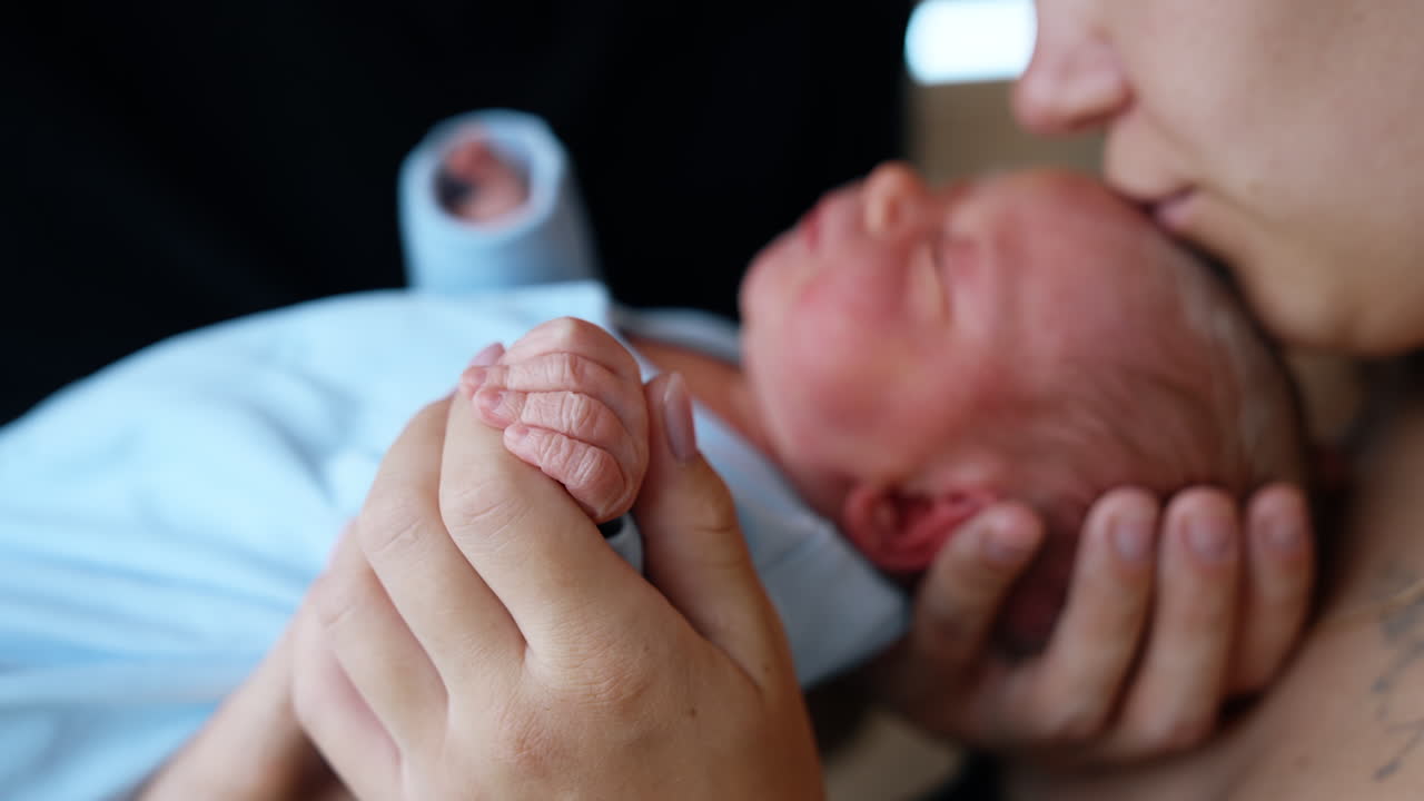 Tiny newborn child wearing blue romper in the parent's hands. Loving mother is holding cute fingers and kissing baby on the head. Close up.