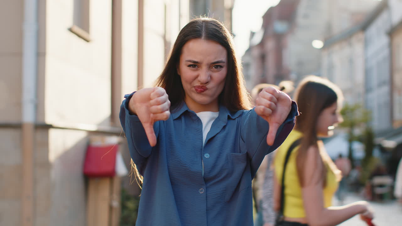 Upset young woman showing thumbs down sign disapproval dissatisfied bad work mistake in city