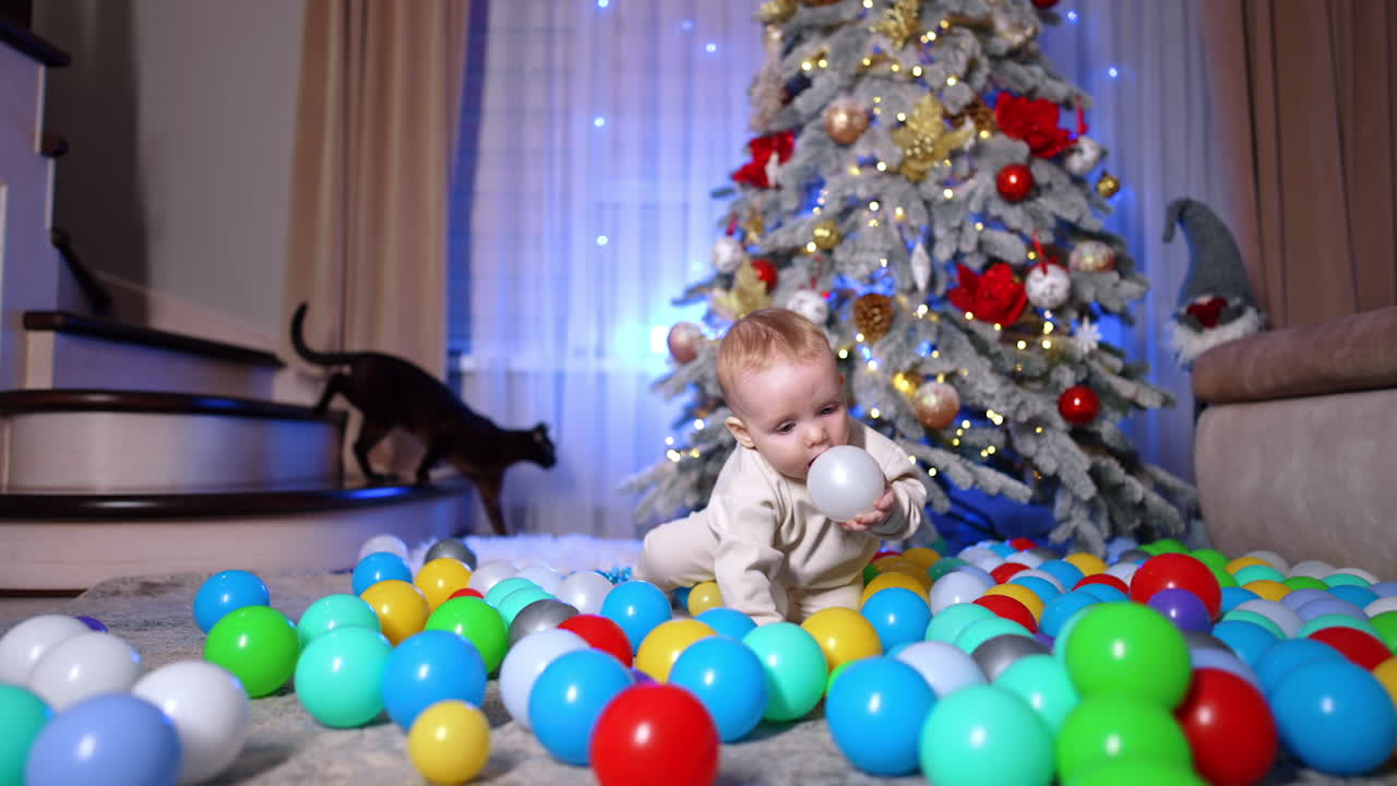 Little baby crawls by the multiple balls scattered on the floor near the Christmas tree. Kid chooses one ball and pulls it to his mouth. Black cat appears at backdrop.