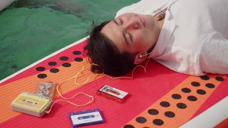 young modern asiatic male sitting on surfboard paddle board while listening to old vintage record tape cassette player with earphone