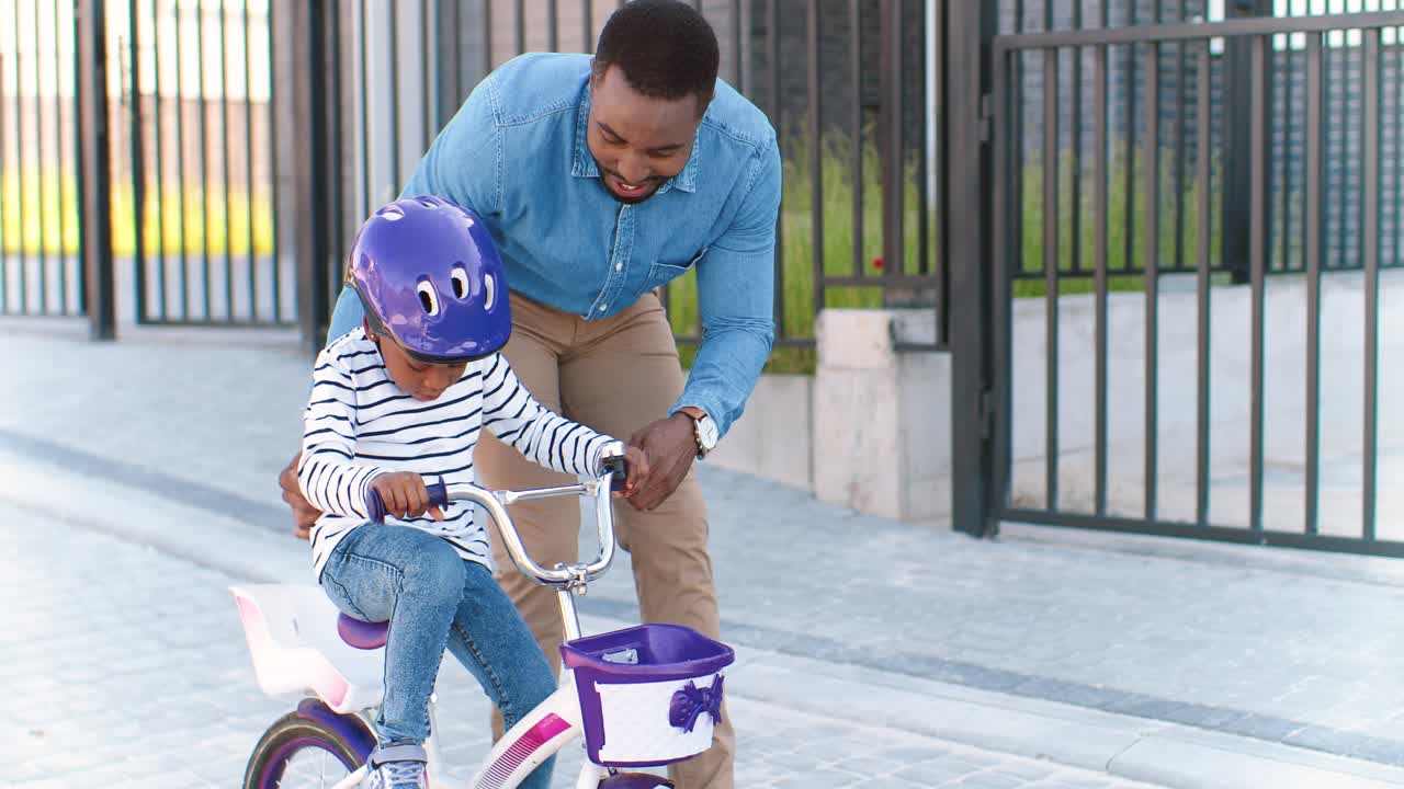 African American father teaching small girl in helmet riding on bike at street in suburb