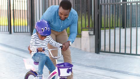 Padre-Afroamericano-Enseñando-A-Una-Niña-Pequeña-Con-Casco-Montando-En-Bicicleta-En-La-Calle-En-El-Suburbio