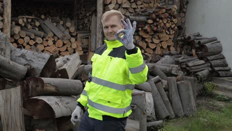 Lumberjack-in-reflective-jacket.-Man-woodcutter-show-ok-sign.-Sawn-logs,-firewood-background