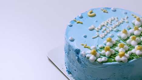 Close-up-shot-of-the-top-of-a-birthday-cake-with-blue-frosting-and-decorated-with-white-and-yellow-frosted-flowers,-with-happy-birthday-greetings-on-top-of-it