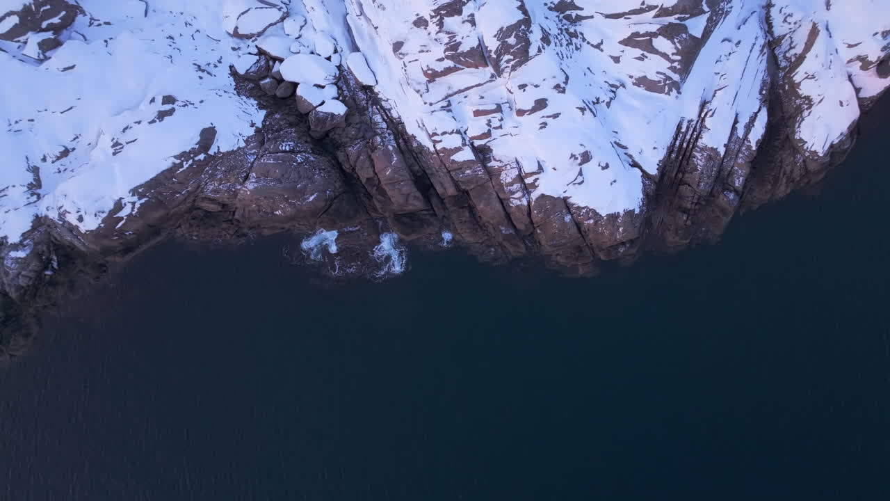 Epic aerial static view of snowy cliff's edge, waves crash on rocks and rugged coastline, Lofoten Islands, Norway
