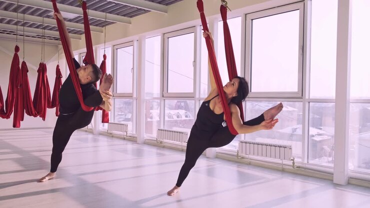 Lezione di yoga aereo: studenti che praticano posizioni di danza con tessuti aerei rossi in uno studio luminoso, migliorando equilibrio e flessibilità per accrescere il benessere fisico.