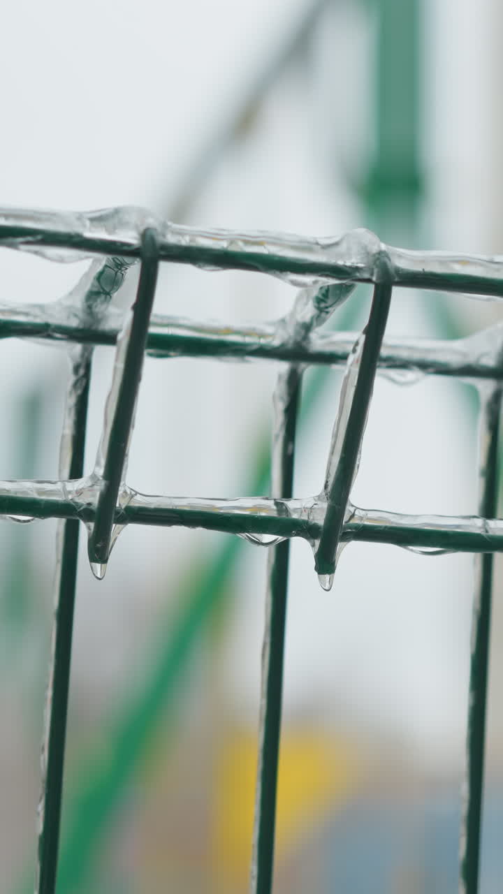 primer plano de una valla metálica verde cubierta de hielo con hielo aferrado a los cables, en un fondo borroso de un patio de recreo nevado, capturando una escena de invierno serena pero fría