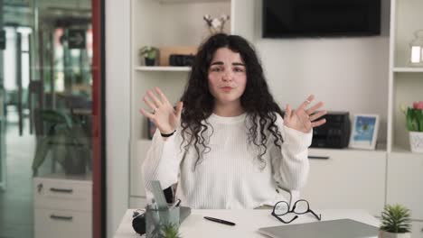 una chica está sentada en una mesa, mirando directamente a la cámara, expresando su satisfacción, gestando emocionalmente con sus manos, tiene el cabello negro rizado, vestida con ropa casual