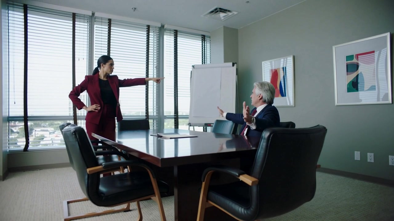 Donna in tailleur bordeaux cambia posizione e indica un uomo seduto a un tavolo da conferenza in un grattacielo.