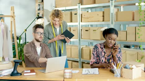 multiethnic designers team typing on tablet, browsing on laptop and talking on the phone and talking to each other in fashion clothing shop