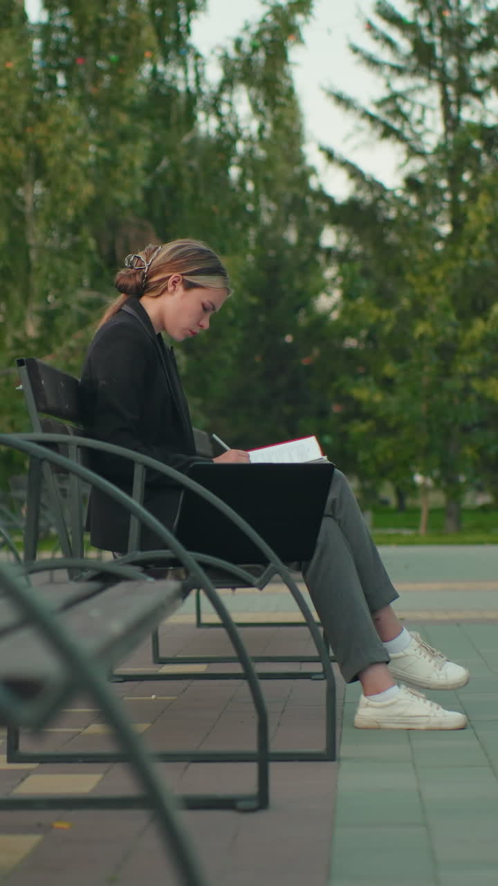 Student in professional attire seated alone on outdoor bench flipping through red folder with pen, laptop beside her, in quiet park with trees and distant people in blur view