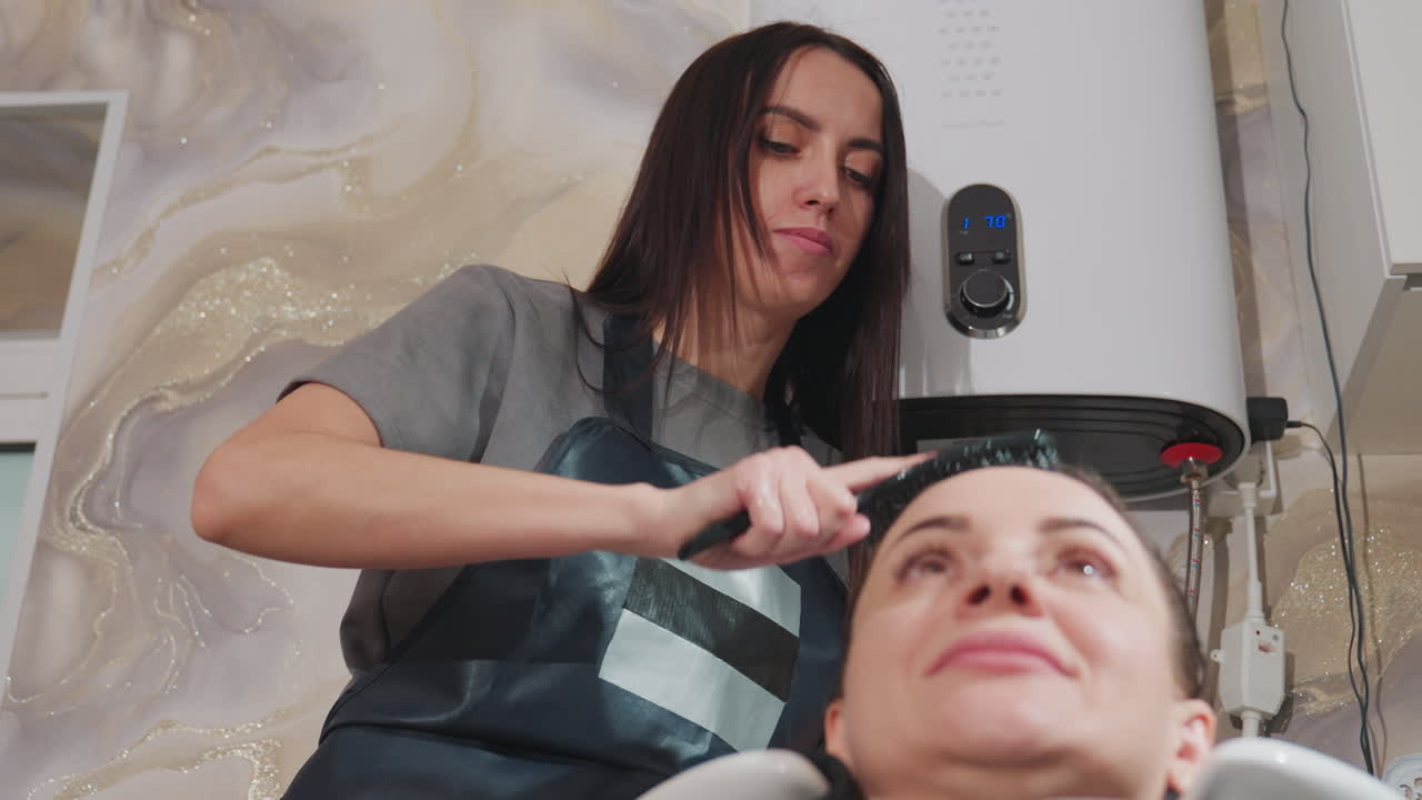 Una clienta blanca en el lavabo de un salón de belleza recibe un suave masaje capilar y un champú de una estilista morena, con limpiador en espuma, sillón reclinable, toalla y capa, iluminación ambiental de spa, cuidado capilar profesional