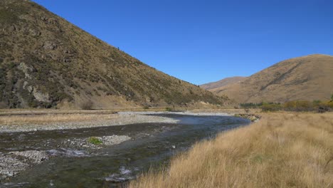 Toma-Panorámica-De-Un-Río-Claro-Que-Fluye-A-Través-De-Colinas-Y-Pastizales-Secos