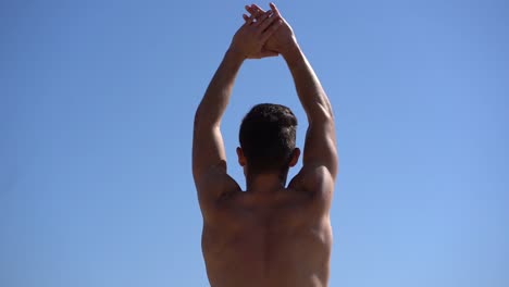 shirtless muscular man stretching arms against blue sky