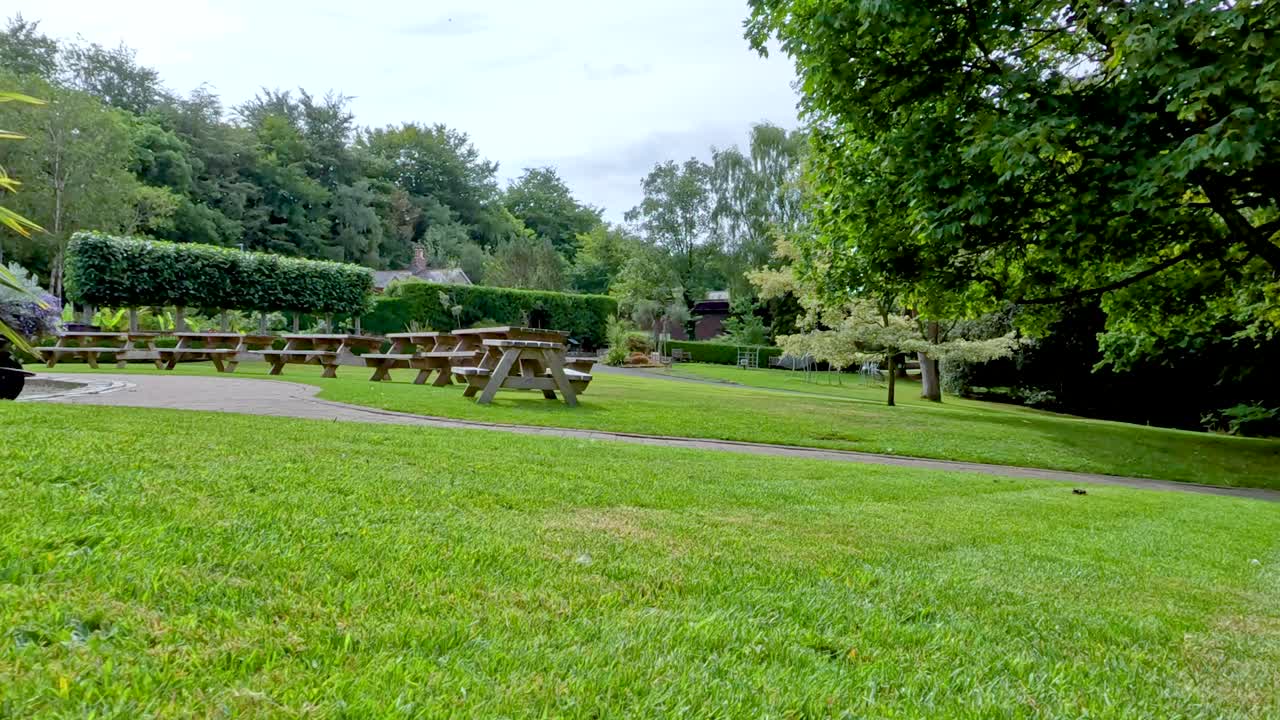 A slow camera pan reveals a lush, green picnic area with benches, large trees, and manicured lawns under bright daylight in Durham, England