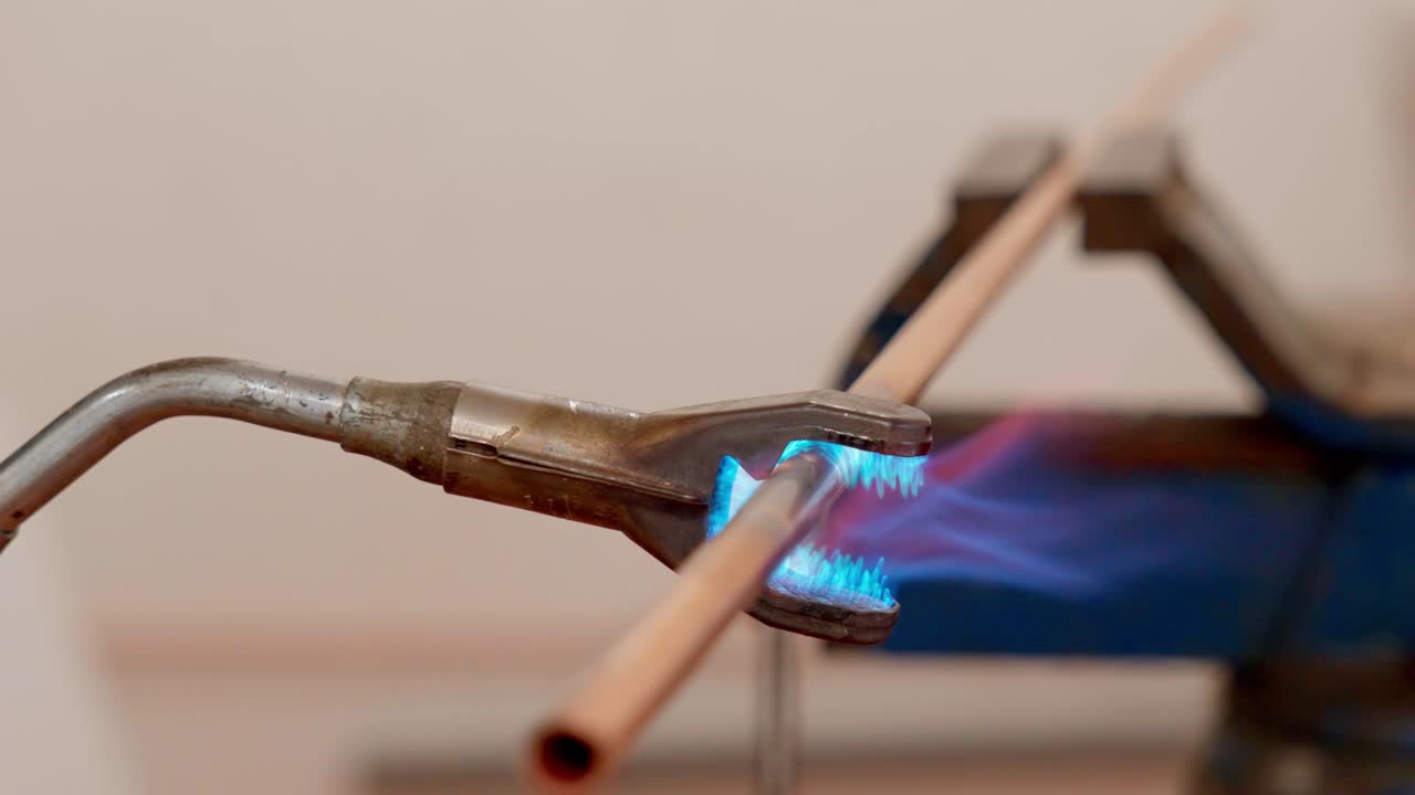 A concentrated blue flame from a torch heats a copper pipe held firmly by pliers for soldering or bending purposes in a workshop setting